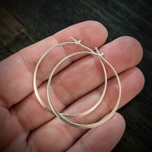 May include: A pair of silver hoop earrings. The earrings are round and have a simple, elegant design. The hoops are held in a hand, and the background is a dark wood surface.
