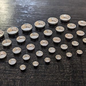 May include: A variety of silver-toned metal bezel settings for crafting jewelry. The bezels are arranged in a staggered pattern on a dark surface.