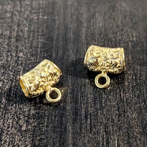May include: Two gold-tone metal cord end caps with an ornate floral design. Each cap has a small loop at the bottom.