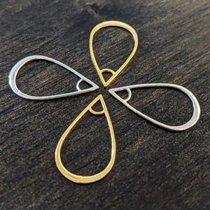 May include: Four teardrop-shaped jewelry findings, two in gold and two in silver, arranged in a flower-like pattern. Each finding has a small, angled loop at the top. The findings are displayed on a dark, textured surface.