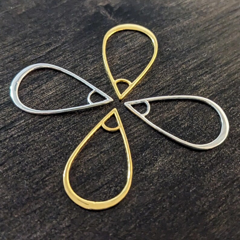 May include: Four teardrop-shaped jewelry findings, two in gold and two in silver, arranged in a flower-like pattern. Each finding has a small, angled loop at the top. The findings are displayed on a dark, textured surface.