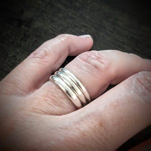 May include: A close-up of a hand wearing four silver rings. The rings are smooth, rounded bands, stacked together on a finger. The rings reflect light, creating highlights. The background is a blurred, dark surface.