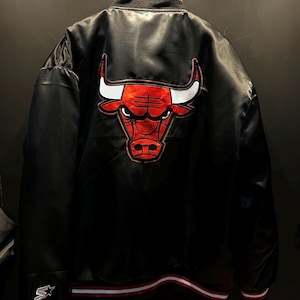 Puede incluir: Chaqueta de satén negro con el logo de los Chicago Bulls en la espalda. El logo es rojo con detalles blancos. La chaqueta tiene cuello y puños a rayas, y un logo de Starter en la manga.