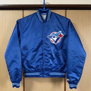 May include: A royal blue satin baseball jacket with a button-up front, featuring a team logo patch on the chest. The jacket has light blue and white striped accents on the collar, cuffs, and hem. The team logo is a blue bird with a baseball and a maple leaf.