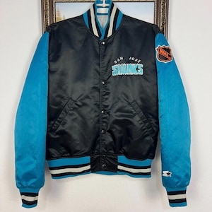 May include: Black satin San Jose Sharks jacket with teal sleeves and accents. Features the team logo and NHL patch. The jacket has a button-up front, pockets, and striped cuffs and hem.