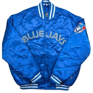 Può includere: Giacca di raso blu con le parole "BLUE JAYS" in bianco sul petto. La giacca ha finiture a righe bianche e blu sul colletto, sui polsini e sull'orlo. Un logo della squadra è sulla manica superiore destra.