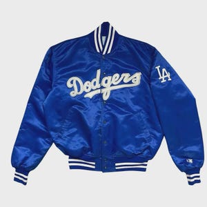 May include: A royal blue satin baseball jacket with the word "Dodgers" in white script across the chest. The left sleeve features a white "LA" logo. The jacket has white and blue striped cuffs and collar.