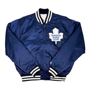 Könnte beinhalten: Eine marineblaue Satinjacke mit weiß-schwarz gestreiften Besätzen an Kragen, Manschetten und Saum. Die Jacke hat ein weißes Ahornblatt-Logo mit dem Text "TORONTO MAPLE LEAFS". Die Jacke hat eine Knopfleiste.