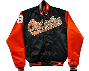 May include: A black and orange satin varsity jacket featuring the word Orioles in orange script across the chest. The jacket has orange sleeves, a striped collar and cuffs, a snap-button front, and the number 8 embroidered on the right sleeve.