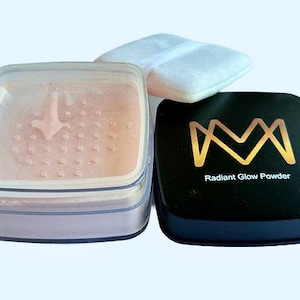 K&ouml;nnte beinhalten: Ein Set von Make-up-Produkten, darunter ein durchscheinender Puder-Kompakt, ein schwarzer Kompakt mit goldfarbenem Schriftzug "Radiant Glow Powder" und ein wei&szlig;es Puderquaste. Das transparente Puder befindet sich in einem transparenten, quadratischen Beh&auml;lter.
