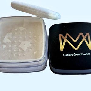 K&ouml;nnte beinhalten: Offener Kompaktpuder Radiant Glow mit einem wei&szlig;en Puderquaste. Der schwarze Deckel des Kompakts hat ein goldenes Logo und die Worte "Radiant Glow Powder". Das Puder hat eine hellbeige Farbe.