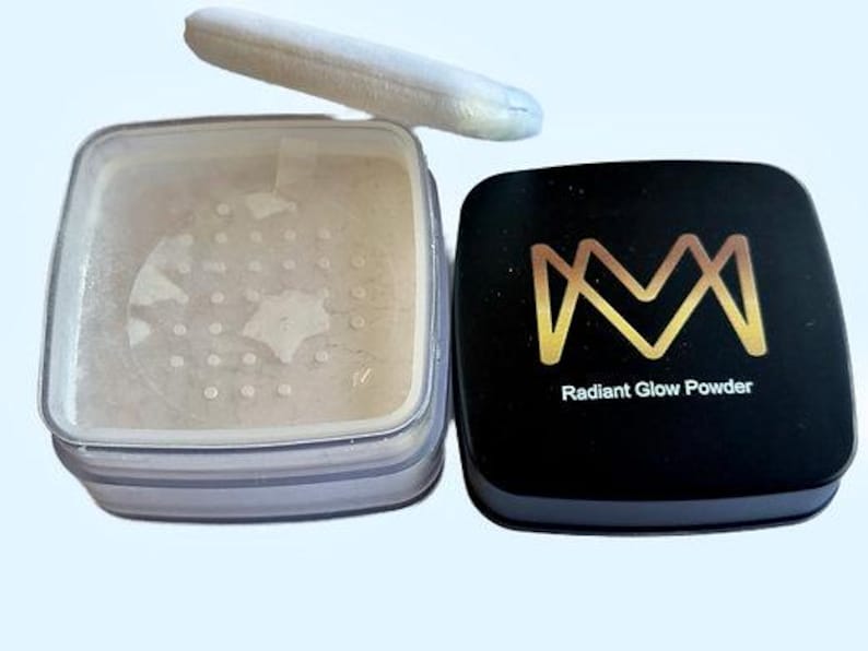 K&ouml;nnte beinhalten: Offener Kompaktpuder Radiant Glow mit einem wei&szlig;en Puderquaste. Der schwarze Deckel des Kompakts hat ein goldenes Logo und die Worte "Radiant Glow Powder". Das Puder hat eine hellbeige Farbe.