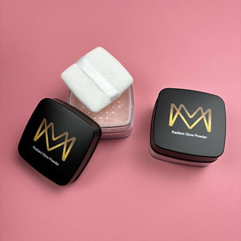 K&ouml;nnte beinhalten: Schwarze quadratische Kompakte mit goldfarbenen Buchstaben und einem wei&szlig;en Puderquaste auf rosa Hintergrund. Die Kompakte tragen den Text "Radiant Glow Powder" und ein stilisiertes "M"-Logo. Der Puderbeh&auml;lter ist transparent.