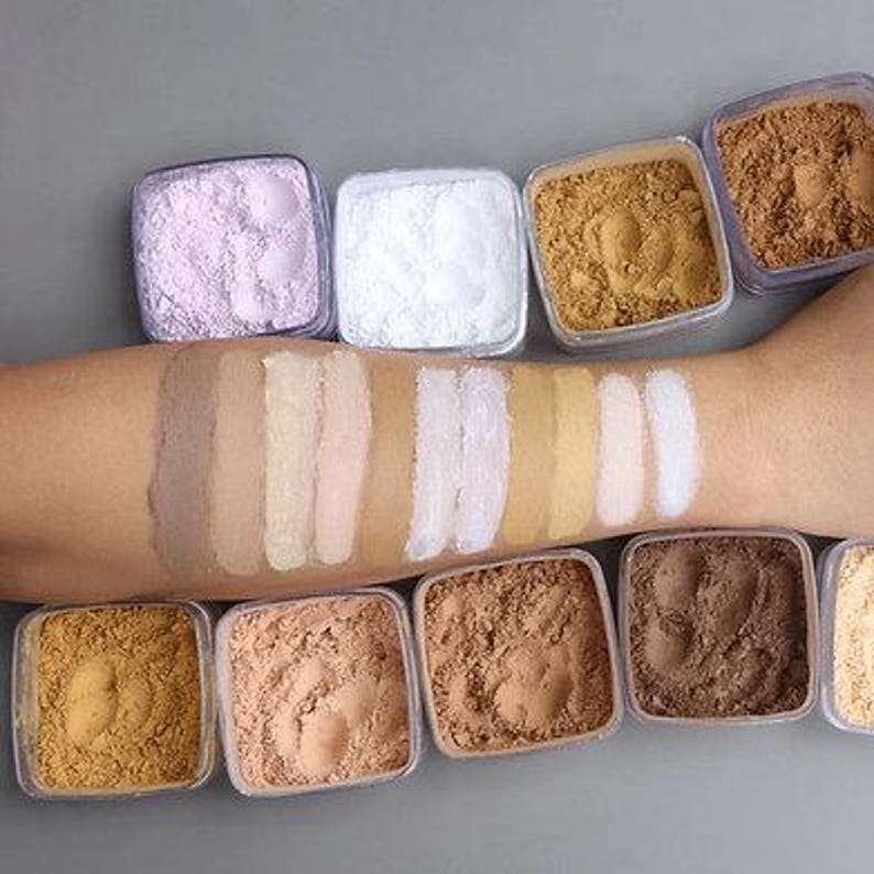 K&ouml;nnte beinhalten: Ein Arm mit Make-up-Farbmustern in verschiedenen Farbt&ouml;nen, von hell bis dunkel, neben kleinen, quadratischen Beh&auml;ltern mit losem Puder in passenden Farben. Die Puder umfassen Farbt&ouml;ne von Wei&szlig;, Beige, Braun und Rosa.