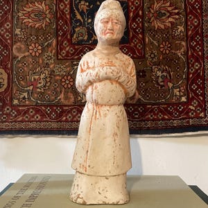 May include: A weathered, off-white ceramic figurine of a person with hands clasped in front. The figure wears a long dress and a hat. The figurine is displayed on a book with the title "Drawings of the Masters French Impressionists".
