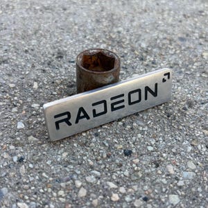 May include: A rectangular silver-toned metal badge featuring the black engraved text RADEON with a small arrow symbol in the top right corner. The badge is positioned against a cylindrical, weathered, and rusted metal object.