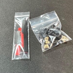 May include: Two clear plastic bags containing hardware components. One bag holds black and red pieces, while the other contains black screws and gold-colored fasteners. The items are arranged on a dark gray surface.