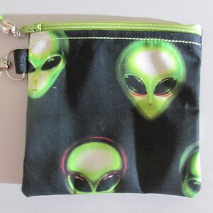 May include: A black zippered pouch featuring a repeating pattern of green alien heads with large dark eyes. The pouch includes a lime green zipper closure, a matching lime green tassel attached to the zipper pull, and a silver metal lobster clasp.