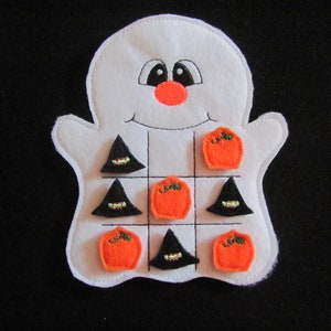 May include: A white felt ghost-shaped Tic-Tac-Toe game. The ghost has a smiling face with black eyes and an orange nose. The game pieces are orange pumpkins and black witch hats. The game is set against a black background.