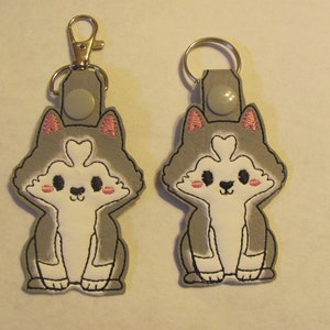 May include: Two embroidered husky keychains. The keychains feature a gray and white husky design with pink accents on the ears and cheeks. One keychain has a silver clasp, and the other has a silver key ring.