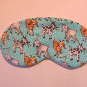 May include: A light blue sleep mask with a repeating pattern of cartoon goats in various colors. The goats are brown, white, and black, with green leaves. The mask has a light blue elastic band.