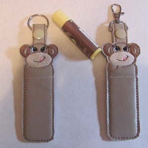 May include: Two brown monkey-shaped keychains with a lip balm holder. Each keychain has a silver ring and a brown button. The lip balm holder is made of brown fabric and has a zipper closure.