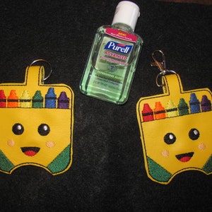 May include: Two yellow crayon-shaped hand sanitizer holders with a smiling face and a green trim. Each holder has a silver key ring and a clear plastic bottle of hand sanitizer inside.