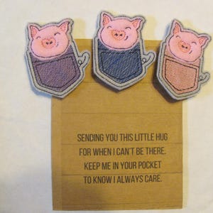 May include: Three pink pig embroidered patches with a pocket design. The patches are on a brown card with the text "SENDING YOU THIS LITTLE HUG FOR WHEN I CAN'T BE THERE. KEEP ME IN YOUR POCKET TO KNOW I ALWAYS CARE."