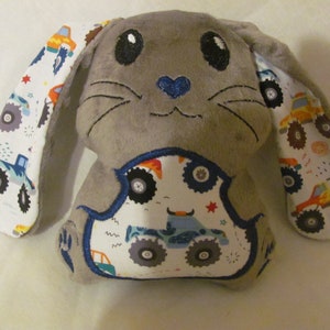 May include: Gray plush bunny with a blue heart nose and embroidered eyes. The bunny's ears are lined with white fabric featuring a pattern of monster trucks in various colors.