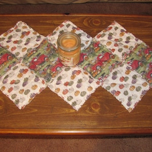 May include: A table runner made of patchwork fabric with a fall theme. The runner is made of squares of fabric with different patterns, including pumpkins, gourds, and trucks. A clear glass jar with a candle is in the center of the runner.
