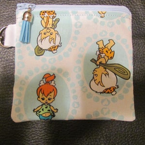 May include: A blue zippered pouch with a white background and a repeating pattern of cartoon characters from the Flintstones. The pouch has a blue tassel attached to the zipper.