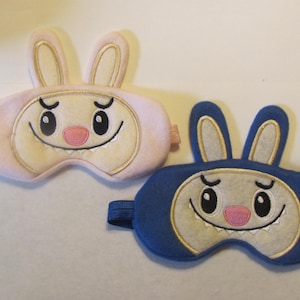 May include: Two sleep masks, one pink and one blue, each designed with a cartoon bunny face. The masks have large eyes, a pink nose, and a smiling mouth. The bunny ears are outlined in gold, and the masks have elastic straps.