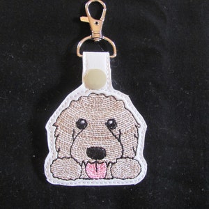 May include: White leather keychain with an embroidered brown and white doodle dog with a pink tongue sticking out.