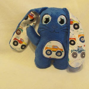 May include: Blue plush bunny with monster truck fabric ears. The bunny has embroidered eyes and a nose. The monster truck fabric features a white background with colorful monster trucks.