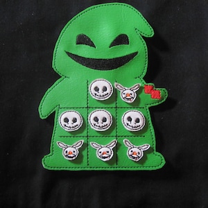 May include: A green Oogie Boogie-shaped tic-tac-toe game. The game board features a grid of nine squares with skull and bunny-shaped game pieces. The Oogie Boogie figure has a stitched design and a smiling face.