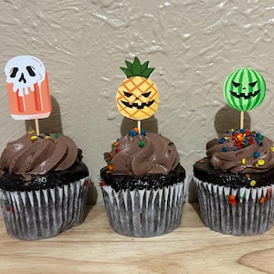 May include: Four chocolate cupcakes with brown frosting and colorful sprinkles, each topped with a decorative paper pick. The picks feature a melting skull popsicle, a jack-o'-lantern pineapple, a jack-o'-lantern watermelon, and a flamingo skeleton.