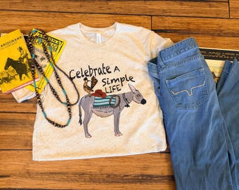 Może przedstawiać: Melanżowy beżowy t-shirt z napisem Celebrate A Simple Life, grafiką osła z czerwonym kapeluszem i turkusowym kocem, zestawiony z niebieskimi dżinsami, długim naszyjnikiem z koralików oraz dwiema starymi książkami w żółtych okładkach.
