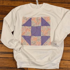 May include: A white crewneck sweatshirt featuring a square quilted patch on the chest. The patch displays a geometric pattern of solid purple squares and triangles alternating with a floral fabric in shades of pink, orange, and blue.