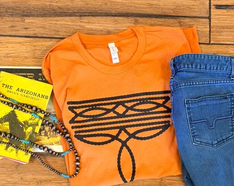 Może przedstawiać: Pomarańczowy t-shirt z krótkim rękawem z czarnym wzorem graficznym przedstawiającym połączone owalne kształty. Koszulka znajduje się obok żółtej książki pod tytułem The Arizonans autorstwa Briana Garfielda, naszyjnika z koralików z turkusowymi akcentami oraz niebieskich dżinsów.