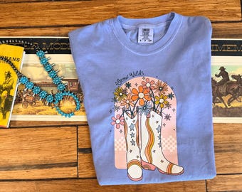 Może przedstawiać: Jasnofioletowy t-shirt z krótkim rękawem z nadrukiem graficznym przedstawiającym dwa białe kowbojskie buty w pomarańczowe i żółte paski z akcentami w kształcie gwiazdek. Bukiet kolorowych kwiatów wyrasta z butów nad różowym łukiem i napisem Grow Wild.