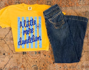 Może przedstawiać: Jaskrawożółty t-shirt z krótkim rękawem z grafiką w jasnoniebieskie pionowe paski, nasionami mniszka lekarskiego i napisem A little more dandelion ciemnoniebieską kursywą, w zestawie z ciemnoniebieskimi dżinsami z tylnymi kieszeniami.