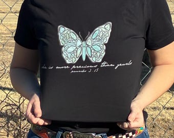 Może przedstawiać: Czarny t-shirt z jasnoniebieskim motylem i szczegółowym wzorem skrzydeł. Koszulka zawiera napis "she is more precious than jewels" i "proverbs 3.15" w białym skrypcie. Koszulka jest połączona z niebieskimi dżinsami i ozdobnym paskiem.