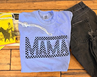Może przedstawiać: Jasnoniebieski t-shirt z krótkim rękawem z napisem MAMA nadrukowanym w czarno-białą szachownicę na klatce piersiowej. Koszulka jest pokazana obok czarnych dżinsów i żółtej książki z ilustracją konia na drewnianej powierzchni.