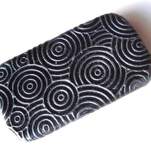 May include: A rectangular, black decorative stone with a pattern of concentric circles. The circles are outlined in a lighter color, creating a textured, eye-catching design. The stone is set against a white background.