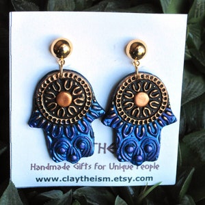 May include: A pair of blue and gold hamsa hand earrings with a gold circle and a sun design in the center. The earrings are handmade and have a gold hook.