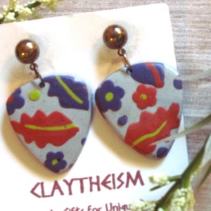 May include: A pair of handmade polymer clay earrings with a white base and colorful floral and leaf patterns. The earrings are shaped like guitar picks and have a silver hook closure. The earrings are displayed on a white card with the text "CLAYTHEISM" printed on it.