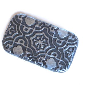 May include: A rectangular, silver-colored metal object with a detailed, embossed pattern. The design features a repeating floral or geometric motif. The object is set against a white background, highlighting its intricate texture and shape.