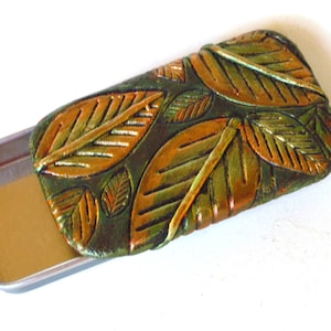 May include: A rectangular metal tin with a decorative lid. The lid is adorned with a leaf design in shades of green, gold, and brown. The tin is open, revealing its empty interior.