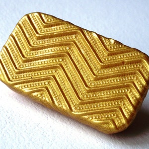 May include: A gold-colored rectangular stamp with a chevron pattern. The stamp has a textured surface with small raised dots.
