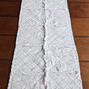 May include: A rectangular, quilted table runner. The runner features a patchwork design with alternating squares of pink floral and plaid fabric. The edges are finished with a ruffled trim. The runner is approximately 60 inches long.